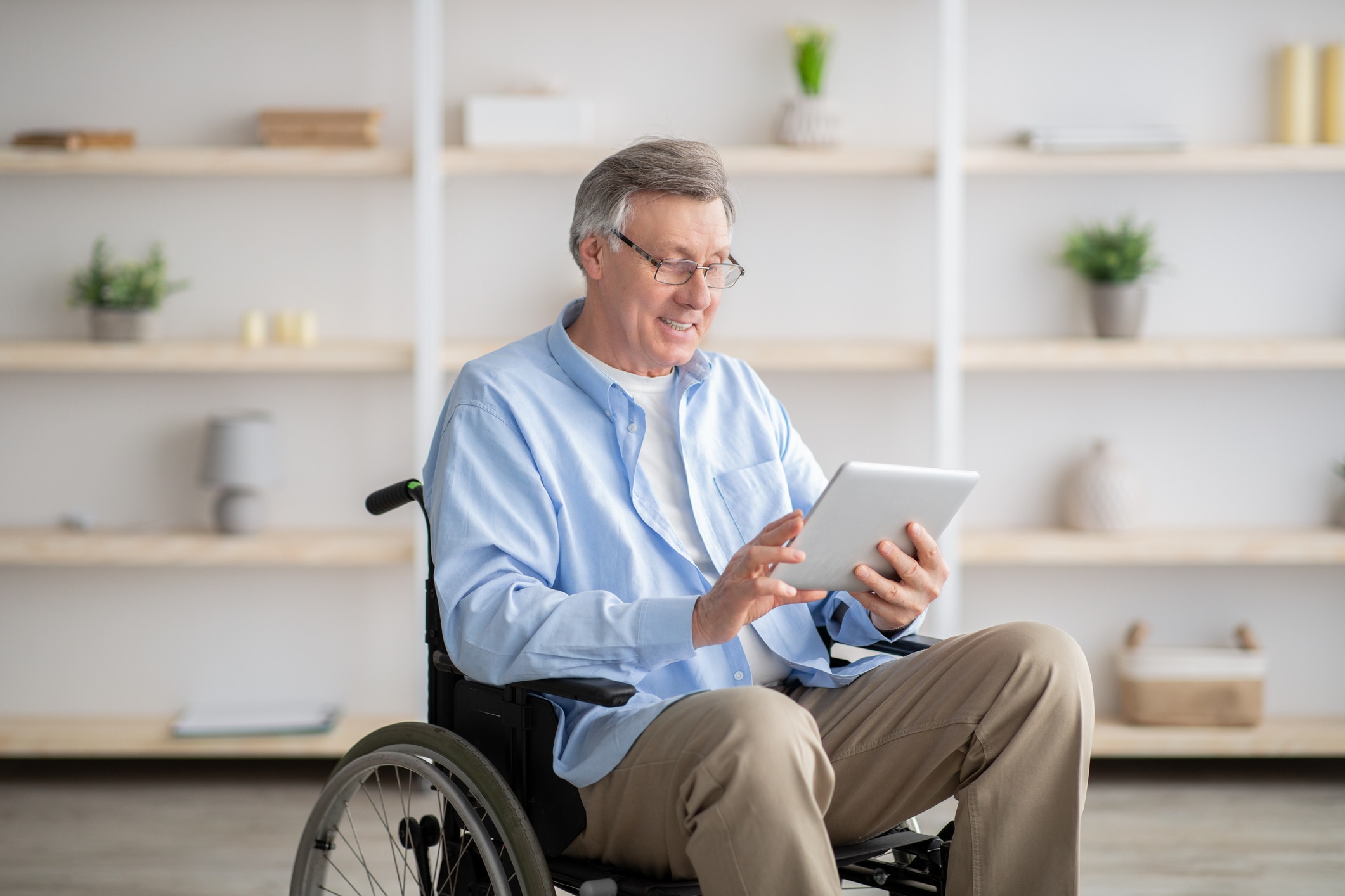 Joyful impaired senior man in wheelchair using tablet computer at home. Positive retired male with physical disability browsing web on touch pad, watching video online indoors