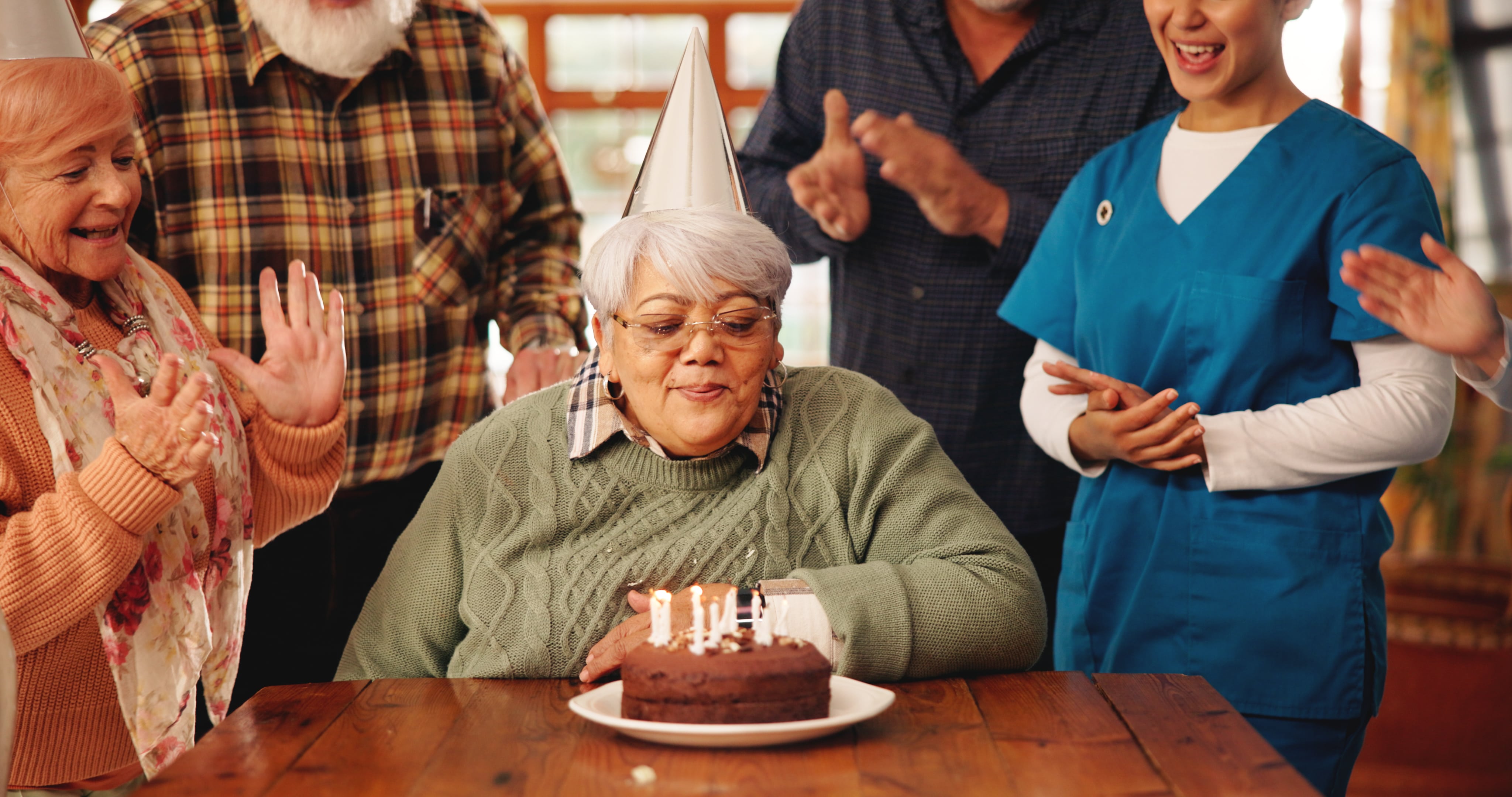 A resident blows out the candles on her birthday cake in a nursing home with friends and medical caregiver